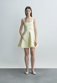 Vrouw in een mouwloze, lichtgele A-lijn jurk met subtiele witte patronen, staand op een grijze vloer tegen een lichtgrijze muur, met witte hakken aan.