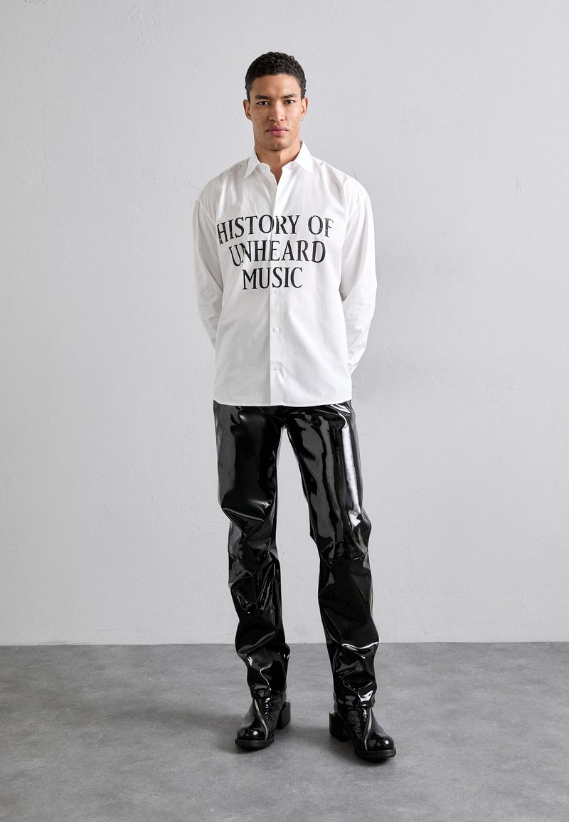 Witte button-up shirt met zwarte tekst "HISTORIE VAN ONGEHOORDE MUZIEK" gecombineerd met glanzende zwarte vinylbroeken en zwarte enkelboots.