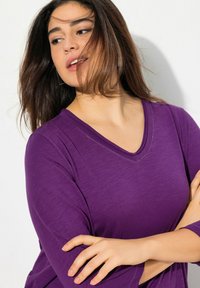 Lila V-Ausschnitt-Oberteil mit 3/4-Ärmeln, weichem Material und dezenten Nähdetails. Das Model posiert mit verschränkten Armen.