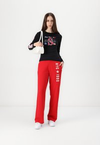 Svart langermet topp med grafisk trykk og røde joggebukser med hvit tekst og stjernesymboler. Kombinert med hvite joggesko og en liten veske.