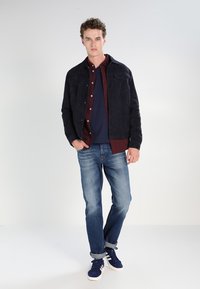 Dark blue suede jacket over a maroon shirt and navy t-shirt, paired with light blue jeans and dark sneakers. Simple design, casual fit.