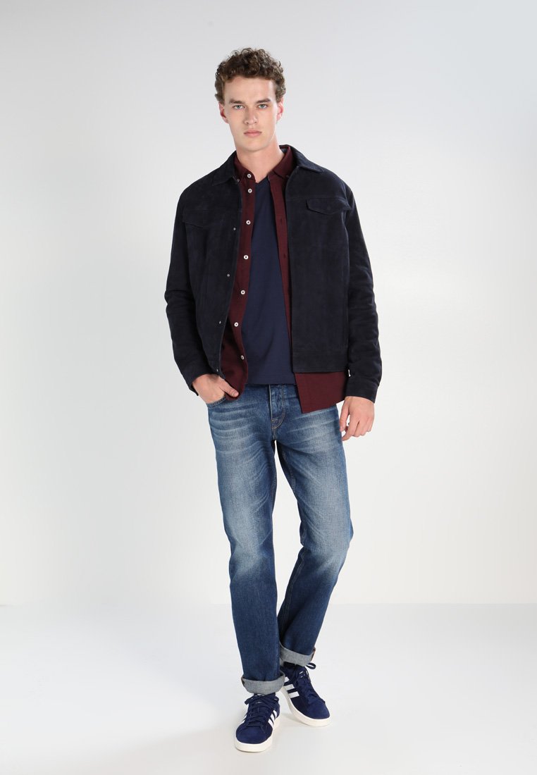 Dunkelblaue Wildlederjacke über einem kastanienbraunen Hemd und einem marineblauen T-Shirt, kombiniert mit hellblauen Jeans und dunklen Sneakers. Einfaches Design, lässige Passform.