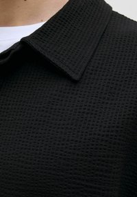 Tissu noir texturé avec un motif en grille, présentant un col pointu. Vue en gros plan du décolleté et des détails du col.