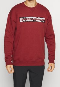Uomo che indossa una felpa maroon di Under Armour con un logo a motivo camo sul petto e pantaloni neri.