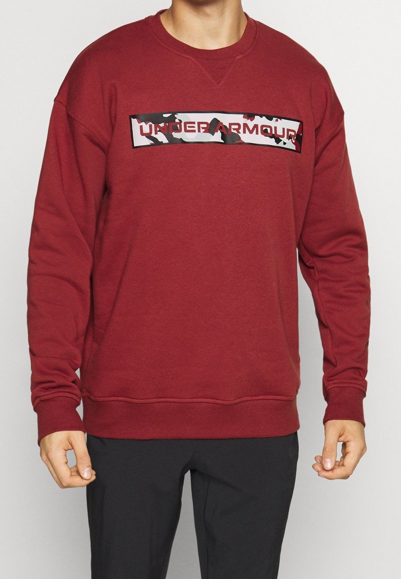 Uomo che indossa una felpa maroon di Under Armour con un logo a motivo camo sul petto e pantaloni neri.