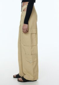 Beige wijde cargo-broek gemaakt van lichtgewicht stof, met grote zijzakken en een zachte textuur. gedragen met zwarte sandalen.