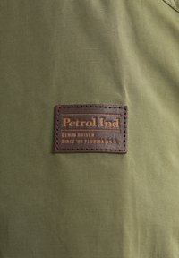 Material textil verde cu o aplicatie din piele. Aplicatia are un text embosat: "Petrol Ind, Denim Driven, Since '89 Florida U.S.A.".