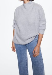 Maglione grigio chiaro a coste con colletto alto e polsini arrotolati, abbinato a jeans a gamba larga leggermente sbiaditi di colore blu.