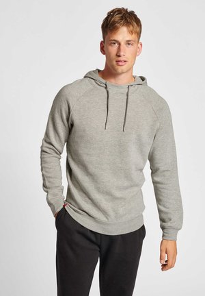 Junger Mann trägt ein hellgraues Kapuzensweatshirt und schwarze Hose, steht mit einer Hand in der Tasche vor einfachem Hintergrund.