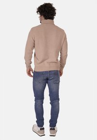 Maglione a collo alto lavorato a maglia beige con texture a costine, indossato sopra jeans di denim blu, visto da dietro, abbinato a sneakers bianche e grigie.