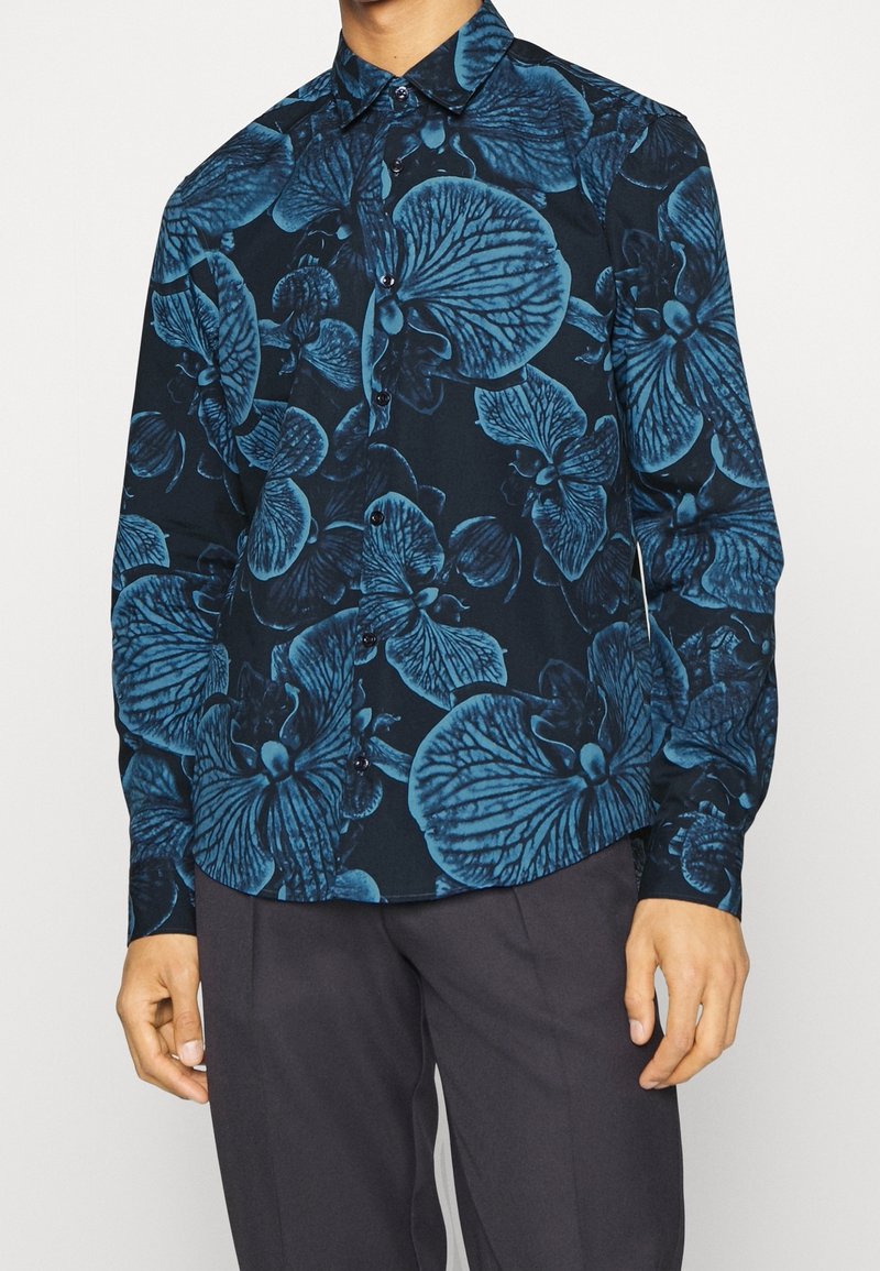 Heren overhemd met lange mouwen van donkere stof met een blauw bloemmotief. Het heeft een knoopsluiting en een puntige kraag.