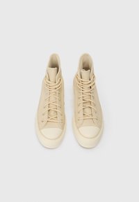 Pár béžových vysokých tenisek s bílými gumovými špičkami a podešvemi, béžovými tkaničkami a zlatými očky, zobrazených zvrchu na bílém pozadí.