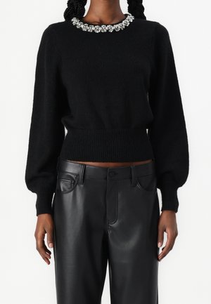 Pull noir duveteux avec encolure ornée d'une fleur bijou, associé à un pantalon en cuir noir taille haute porté par une personne debout.
