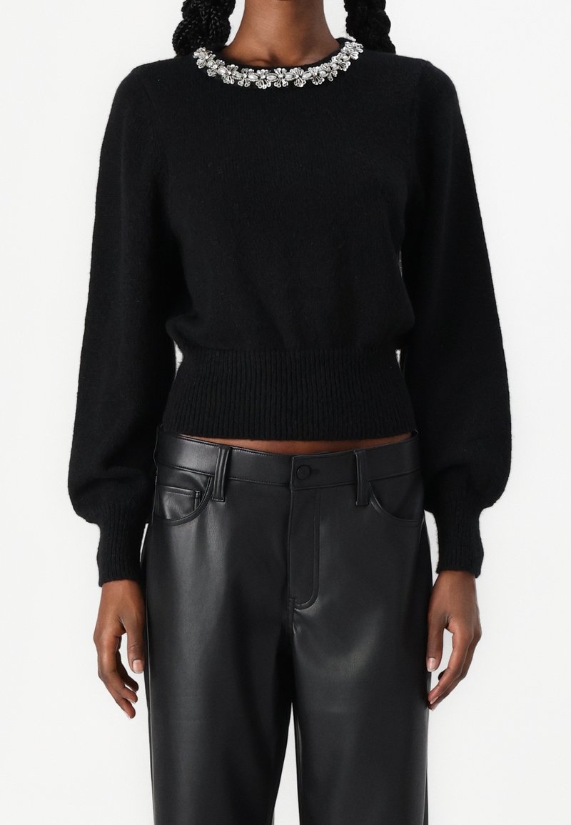 Pull noir duveteux avec encolure ornée d'une fleur bijou, associé à un pantalon en cuir noir taille haute porté par une personne debout.