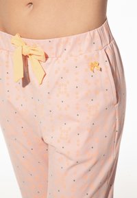 Lichtroze pyjamabroek met oranje bloemen- en groene stippenpatronen, voorzien van een gele strik in de taille en een klein geel strikdetail.