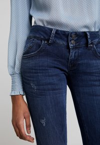 Mörkblå denimjeans med slim fit, som har två framfickor, slitna detaljer och en dubbelknäppning i midjan.