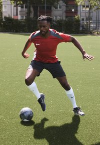 Červený športový dres Nike so sivými akcentmi, čierne kraťasy a biele ponožky. Hráč dribluje čiernu a sivú futbalovú loptu na zelenej tráve.