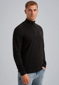 Pull-over noir à col zippé en tissu tricoté lisse, avec des accents côtelés au niveau du col et des poignets, associé à un jean bleu clair.
