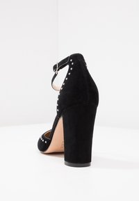 Svart mocka högklackad sko med ankelrem, prydd med silvernitar längs kanten och en tjock klack. Slät textur och klassisk design.
