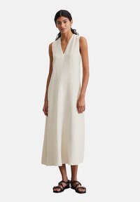 Weißes Leinenkleid ohne Ärmel mit V-Ausschnitt, fließender Silhouette und knielangem Saum. Trägt schwarze Riemchensandalen.