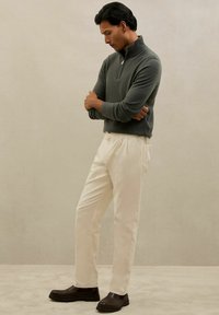 Pelēka zip-up džemperis un krēmkrāsas bikses ar brīvu piegriezumu. Brūni slip-on apavi ar biezu zoli noslēdz apģērbu.