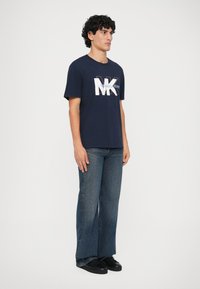 Hombre con cabello rizado que lleva una camiseta de logo de Michael Kors en color marino, unos vaqueros de pierna ancha azules y zapatillas negras, de pie frente a un fondo blanco.
