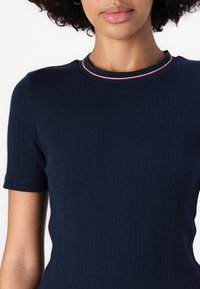Cămașă tricotată rib cromată, de culoare bleumarin, cu guler rotund căptușit cu dantelă roșie și albă. Mâneci scurte și design ajustat, textură netedă.