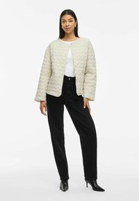 Gequilte crèmekleurige jas met een ronde halslijn, open voorkant en lange mouwen, gecombineerd met zwarte high-waisted broek en zwarte puntige hakken.