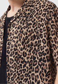 Homme portant une chemise à manches courtes à boutons avec un imprimé léopard marron et noir par-dessus un t-shirt noir.