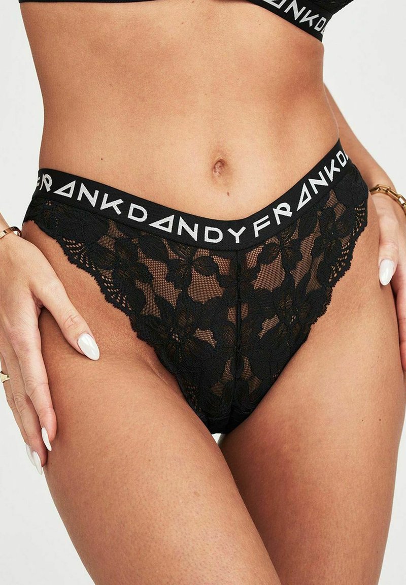 Tanga in pizzo nero con un motivo floreale, una morbida cintura elastica con branding stampato e bordi scallopati lungo le aperture delle gambe.