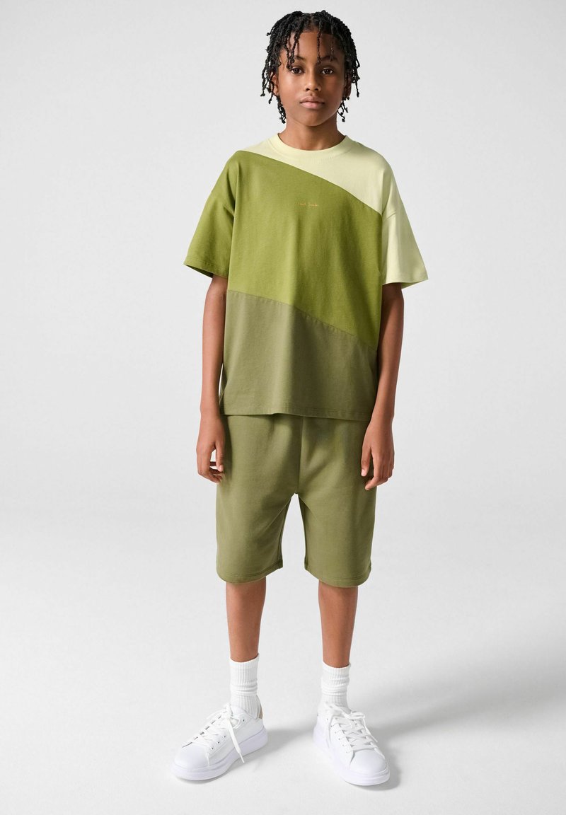 Barn med flettet hår iført en t-shirt i grøn og beige farveblok, grønne knælange shorts, hvide sokker og hvide sneakers, stående på en grå gulv.