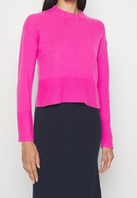 Fuksjowy krótki sweter z długimi rękawami i falbanianym dekoltem. Miękka faktura i dopasowany fason. Połączony z prążkowaną, ciemną spódnicą.