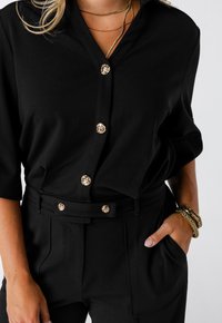 Vrouw in zwarte blouse met knopen en bijpassende broek met gouden knopen, gouden armbanden en gelaagde gouden kettingen, hand in de zak.