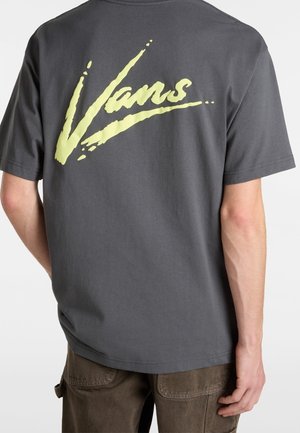Rückenansicht einer Person, die ein dunkelgraues Vans-T-Shirt mit großem gelbem Vans-Logo über dem oberen Rücken und braunen Hosen trägt.