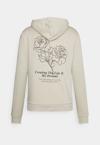 Beige hættetrøje med sorte linjetegnede roser på ryggen og tekst, der lyder "Creating The Life Of My Dreams."