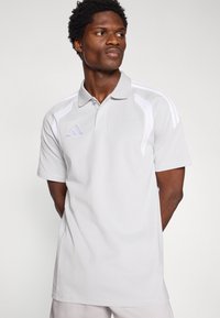 Ανοιχτό γκρι πόλο μπλούζα με λευκές λεπτομέρειες, κοντά μανίκια, σχέδιο με γιακά, με το λογότυπο της Adidas σε μωβ στο αριστερό μέρος του στήθους.