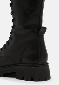Bota de combate de cuero negro con cordones en la parte frontal, suela de goma gruesa con tacos y un pequeño detalle de logo en el lado.