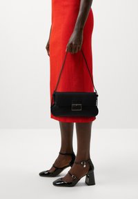 Sac à main en cuir noir de forme rectangulaire, avec un fermoir à l'avant et une bandoulière. Porté avec une robe texturée rouge vif et des chaussures noires.