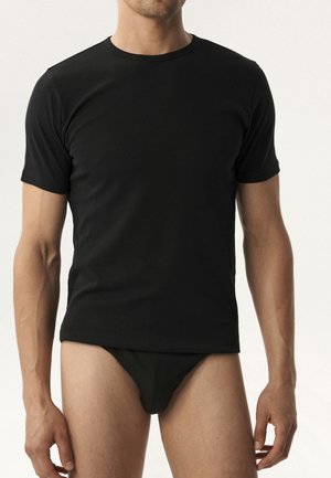 Mężczyzna ubrany w prosty, czarny t-shirt z krótkim rękawem i okrągłym dekoltem oraz czarne bokserki na jasnym tle.