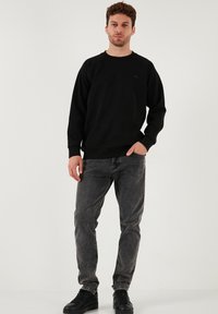 Črna dukserica s krožnim ovratnikom in rebrastimi manšetami, v kombinaciji s temno sivo jeans hlačami in črnimi športnimi copati. Tkanina izgleda mehka in strukturirana.