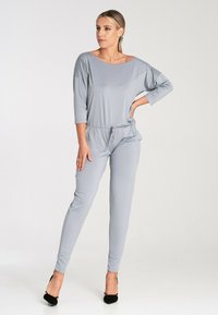 Jasnoszary kombinezon o luźnym kroju, z szerokim dekoltem w łódkę i rękawami o długości trzech czwartych. Posiada regulowany pas oraz zwężane nogawki.