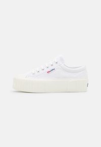 Baskets en toile blanches avec une semelle épaisse texturée, arborant un petit logo rouge et bleu sur le côté et six œillets pour les lacets.