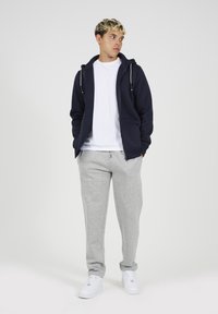 Marine blauwe rits hoodie, witte t-shirt, grijze joggingbroek en witte sneakers. Casual fit, gestructureerde stof met trekkoordaccenten op de hoodie.