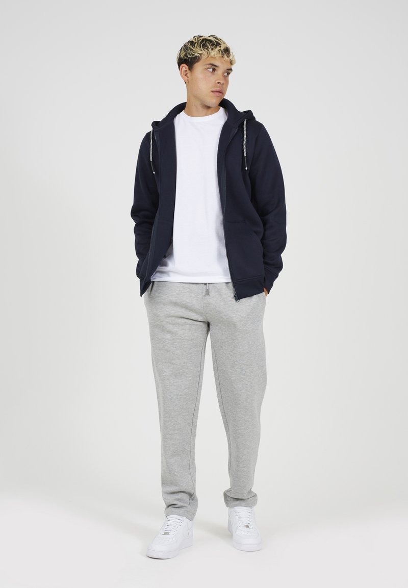 Marine blauwe rits hoodie, witte t-shirt, grijze joggingbroek en witte sneakers. Casual fit, gestructureerde stof met trekkoordaccenten op de hoodie.
