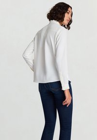 Weiße Bluse mit Rüschenkragen und -manschetten, lange Ärmel, glatte Textur, kombiniert mit dunkelblauen, eng anliegenden Jeans, Ansicht von hinten.