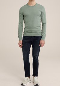 Světle zelený svetr s kulatým výstřihem, přiléhavými rukávy a žebrovanými manžetami, spárovaný s tmavými džíny a bílými sportovními botami.