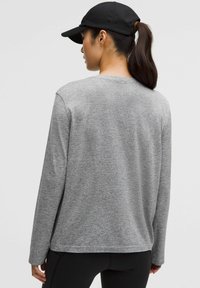 Maglietta grigia a maniche lunghe con tessuto testurizzato, scollo rotondo e vestibilità rilassata; abbinata a leggings neri e un cappellino nero.