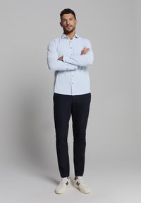 Lichtblauw overhemd met lange mouwen, donkere broek en witte sneakers met blauw logo. Het overhemd heeft knopen en een klassieke kraag.
