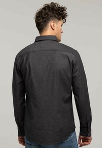 Mężczyzna z kręconymi włosami, ubrany w ciemnoszary, długi rękaw koszulę i niebieskie dżinsy, pokazany z tyłu na jednolitym tle.