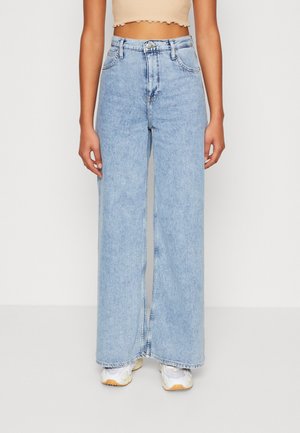 Person iført lyseblå, højtaljede bukser med vide ben, beige crop top og hvide sneakers, stående mod en hvid baggrund.